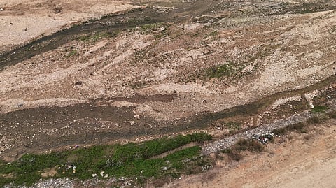 Aerial view of a dry, rocky riverbed with minimal green vegetation. Sparse patches of grass and shrubs appear along the banks, conveying arid conditions.