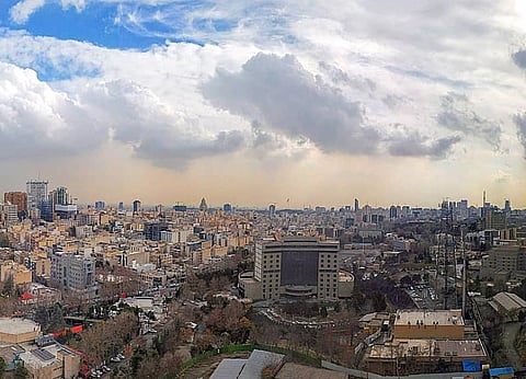 Panoramic view of a sprawling cityscape under a partly cloudy sky. Tall buildings and dense housing dominate the scene, conveying urban hustle.