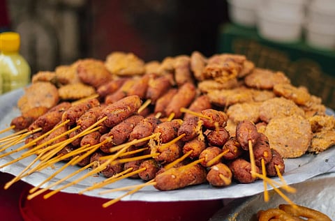 A street food display featuring skewered sausages and fried patties on a metal tray, set against a street market backdrop. The scene exudes a casual, inviting vibe.