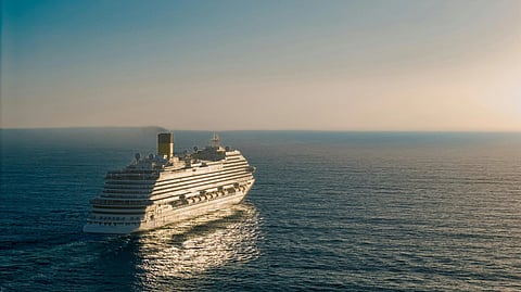 A large cruise ship sails across the calm ocean under a clear sky, with sunlight reflecting off the water, creating a serene and peaceful atmosphere.