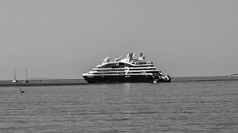Black and white photo of a serene beach scene, with two people sitting on pebbles and one swimming in the calm sea. A cruise ship is visible on the horizon.