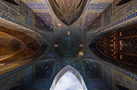 Intricate blue and gold mosaic ceiling of a mosque, with geometric patterns and archways. The view follows the architectural symmetry, evoking awe and reverence.