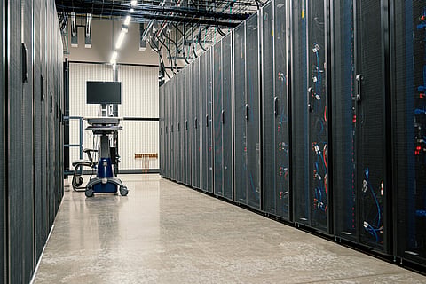 A modern data center aisle with rows of server racks on both sides. A computer on a wheeled cart stands on the polished floor, under bright ceiling lights.