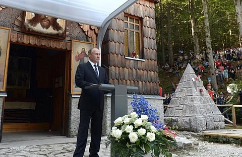 A man in a suit speaks at a podium outside a rustic chapel with religious icons. A flower arrangement and small crowd in a forest setting are visible.
