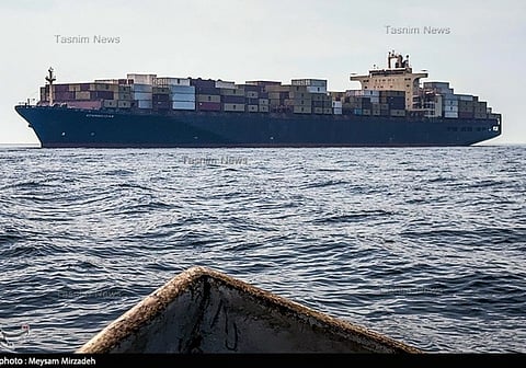 A large cargo ship loaded with colorful shipping containers sails on a calm sea under a clear sky. The bow of a smaller boat is visible in the foreground.
