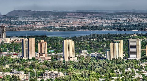 A cityscape with tall skyscrapers amidst lush greenery, set against a wide river and distant mountains under a clear sky, conveying tranquility.
