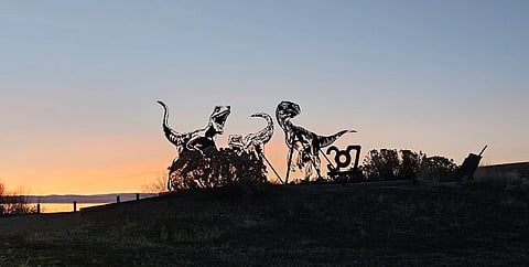 Silhouette of dinosaur sculptures at sunset, on a hill with a vast sky. The scene is tranquil, with warm hues and a clear horizon.