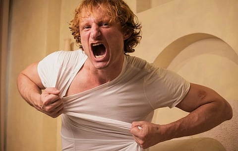 A man with curly hair is shouting angrily while pulling at his white T-shirt. The background is neutral, adding focus to his intense emotion.
