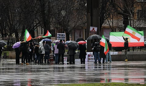 A group of people gathers in a rainy square, holding Bulgarian and Iranian flags. Umbrellas shield them, reflecting a somber protest atmosphere.