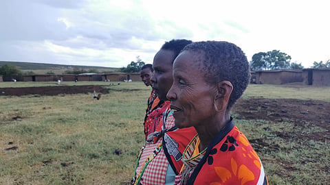 Three women in vibrant, traditional attire stand in a grassy field, facing right. The sky is overcast, with huts and trees in the background.