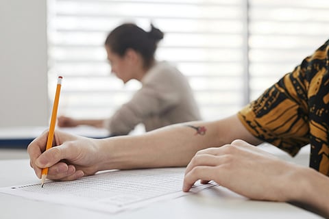 A student in a yellow patterned shirt writes with a pencil on a test paper, focusing intently. Another student is blurred in the background, creating a studious atmosphere.