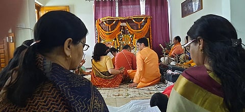 A group of people in traditional attire are gathered indoors for a religious ceremony. They sit on a rug facing an ornate altar adorned with marigold garlands. The atmosphere is solemn and focused.