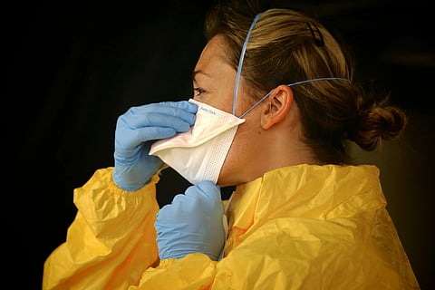 A healthcare worker in a yellow protective gown, blue gloves, and a white face mask adjusts her mask. The setting is serious and focused.