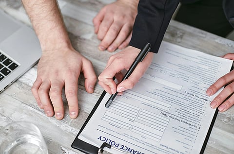Hands signing an insurance policy document on a clipboard, with a laptop nearby on a wooden table. The scene conveys a formal, professional tone.