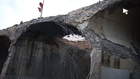 A partially collapsed concrete structure with exposed rebar, showing significant damage. A flag waves atop, suggesting resilience amid destruction.