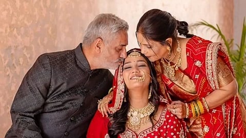 Twisha Sharma in a red and gold lehenga sits between her parents, smiling joyfully. Her father kisses her forehead, while her mother leans in affectionately.