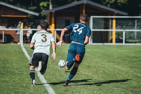 Two soccer players sprint towards the ball on a sunny field. One wears a white jersey and the other blue, conveying a sense of competition and focus.