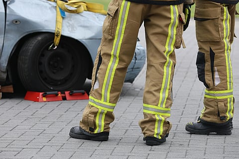 Two firefighters in tan uniforms with reflective stripes stand beside a damaged gray car on a paved surface. Red chocks secure the vehicle's wheel.