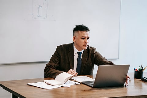 Businessman in a suit working on a laptop at a wooden desk with papers and an open notebook. A whiteboard is in the background, conveying focus.