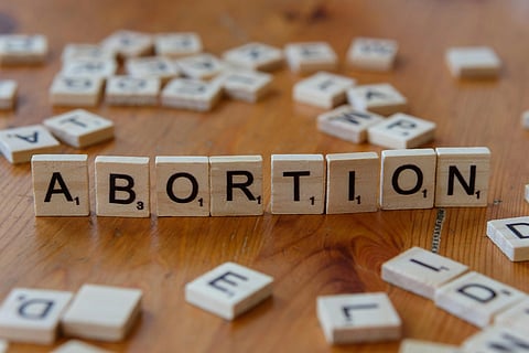 The word "ABORTION" is spelled out with wooden letter tiles on a wooden surface, surrounded by scattered, random tiles, conveying a serious tone