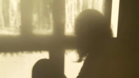 A shadowy silhouette of a person sitting by a window. The image is dimly lit, conveying a serene and contemplative atmosphere.