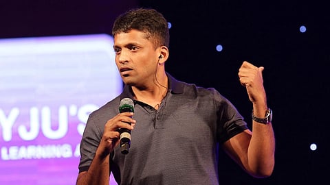 In the image Byjus founder Byju Raveendran  is shown behind him is a screen with half of Byjus logo