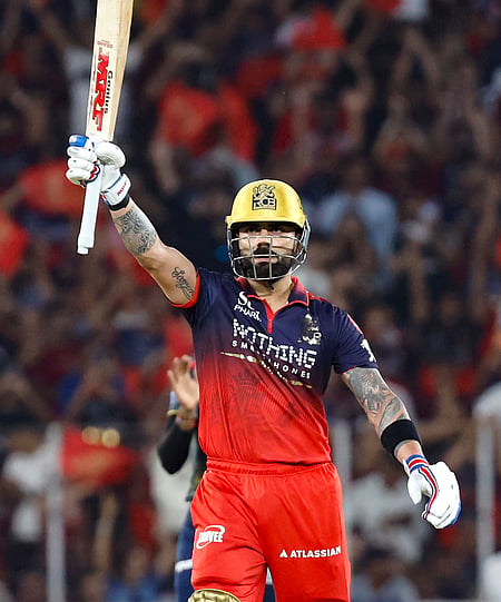 Image of Virat Kohli in a red and blue uniform and yellow helmet raises his bat triumphantly amidst a cheering crowd, showcasing a moment of celebration and achievement.