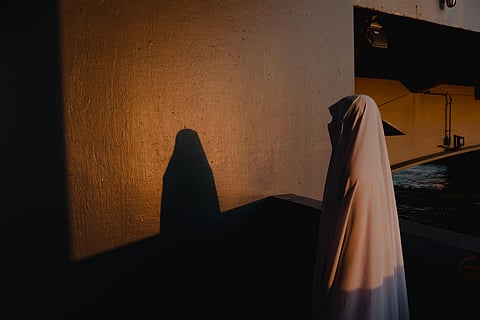 A person in a white robe stands under a bridge, casting a shadow on a sunlit wall. The warm light creates a serene and contemplative mood