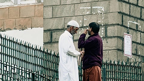 Two men conversing on a sidewalk, one in a white robe and cap, the other in a dark jacket. They stand near a stone building and metal fence, exchanging a serious, engaged expression