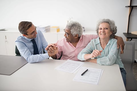 A joyful elderly couple sits at a table with a financial advisor. Papers and a pen are nearby, signifying discussions. The mood is friendly and supportive.