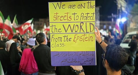 A person holds a sign reading "We are on the streets to protect the world from the Epstein class" in a large protest. Iranian flags are visible, and there is a sense of urgency among the crowd.