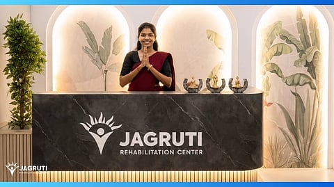 A smiling woman stands behind a modern reception desk at Jagruti Rehabilitation Center, decorated with plants and soft lighting, creating a welcoming atmosphere.