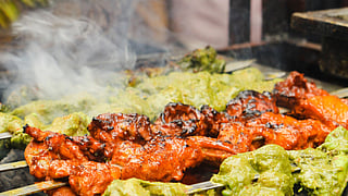 Grilled skewers with green and red marinated meats sizzle over charcoal, emitting steam in a bustling street food setting.