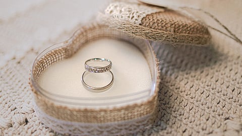 Two silver rings rest in a heart-shaped box with a crocheted edge on a textured fabric. A blurred glass dome covers a plant in the background, evoking a romantic tone.