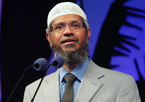 Image of Islamic Preacher Zakir Naik with a beard and glasses speaks into two microphones. He is wearing a white cap, light gray suit, blue shirt, and patterned tie, set against a purple background.