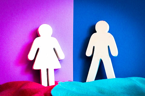 Wooden cutouts of a woman and a man stand against a pink and blue split background. Red and teal fabric drapes below, creating a contrast