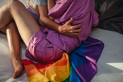 Two people in colorful clothing sit closely on a bed, partially covered by a rainbow flag. The scene feels intimate and supportive