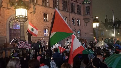 Crowd gathers at night outside a historical building. People hold Palestinian and Lebanese flags. A sign reads “Jews Against Genocide.” The mood is solemn.