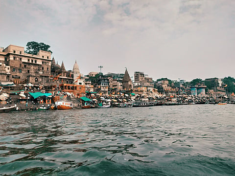Riverside view of Varanasi, India, showing densely packed historic buildings, temples, and busy ghats along the Ganges under a partly cloudy sky.