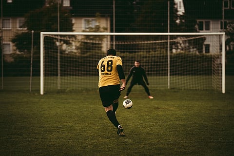 Soccer player in yellow jersey prepares to shoot a penalty, facing the goalkeeper in black, with focus and tension on a green field.