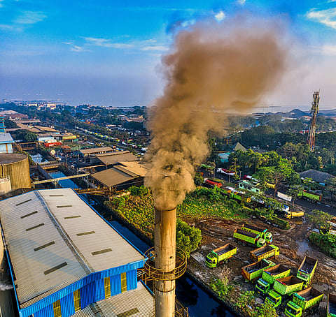 A factory chimney releases thick dark smoke over an industrial area with several buildings, green trucks, and some greenery visible under a blue sky.