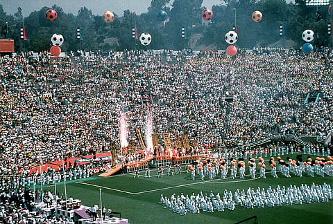 Image of Sports - 1994 World Cup 01 A packed stadium with thousands of spectators and performers creating a festive atmosphere. Giant soccer ball balloons float above, while performers in coordinated outfits carry props on a vibrant field, with colorful smoke and fireworks enhancing the celebratory mood.