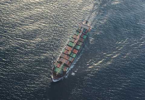 Aerial view of a large cargo ship with multiple containers sailing through the open ocean, creating a white wake. The water is calm, reflecting soft sunlight.