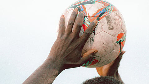 A soccer player in a white jersey prepares to throw the ball, eyes focused. The ball features colorful patterns, set against a clear sky.