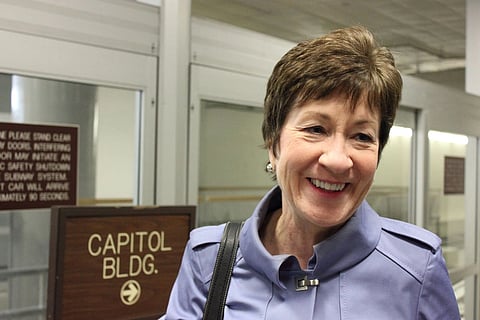 Sen. Susan Collins in a light purple coat stands near a sign that reads "Capitol Bldg." in an indoor setting, conveying a positive tone.