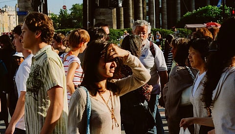 A diverse crowd of people walks on a sunny city street. A woman shields her eyes from the sun. The atmosphere is lively and bustling.