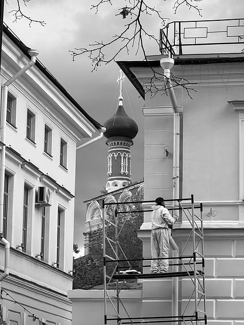 A person in overalls stands on scaffolding, painting a building in a narrow alley. In the background, an ornate, domed church tower rises under a cloudy sky.