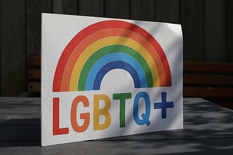 A greeting card featuring a rainbow with "LGBTQ+" in bold, colorful letters, set against a shaded wooden table and a dark fence background.