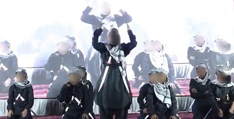 A group of children in uniform and keffiyeh scarves sit and stand on a stage, with blurred faces, conveying a serious tone.