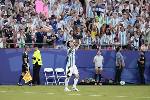Football player Lionel Messi in a blue and white jersey celebrates with arms raised, facing cheering crowd in a stadium. Emotion is jubilant and triumphant.